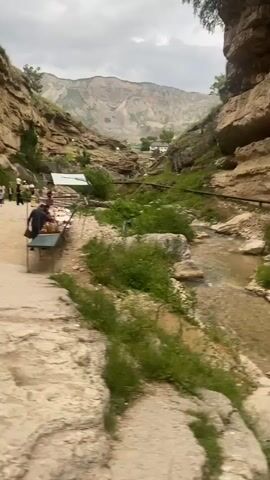 The video pans from a rocky cliff face and a stream to a dirt path. A person wearing a white top and white pants walks towards the camera on the path. Another person wearing a brown shirt and a black skirt walks away from the camera on the same path. In the background, two other people walk away from the camera on the path. The environment consists of a rocky canyon with a stream running through it and green vegetation.