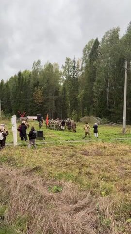 A group of people in vintage military uniforms, some holding flags and what appear to be replica weapons, are gathered in a grassy, wooded area. Two individuals in the center are engaged in a staged interaction, possibly a reenactment. In the foreground, a person is filming the event with a camera, and another person is observing. A large, green, tank-like object is visible in the background, as are trees and a cloudy sky.