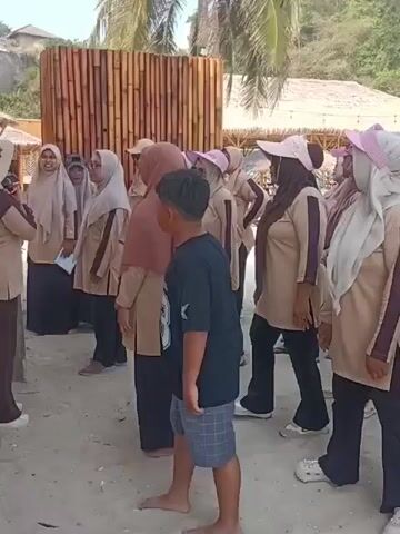 A group of women, some wearing hijabs and light pink visors, are gathered on a sandy beach. They are dressed in matching tan uniforms with darker accents. A young boy in a black t-shirt and plaid shorts stands among them, facing away from the camera. Behind the group is a bamboo structure and thatched-roof cabanas, with a hill and palm trees in the background. The scene appears to be a group outing or activity.