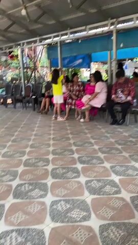 The video shows an indoor gathering with a corrugated metal roof. Several people are seated on chairs arranged in a row. The floor is tiled with a patterned design. In the foreground, a young girl in a yellow top and white shorts stands with her hands near her head. To her right, a woman in a pink dress is seated, and next to her, a man in a patterned shirt is looking down at a device. Other individuals are visible in the background, some seated and others standing near a table covered with a white cloth. The overall atmosphere appears to be a casual event.