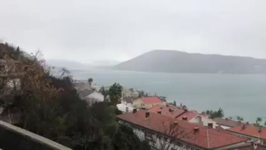 The video shows a coastal scene in what appears to be Montenegro during a heavy rain. In the foreground, there are rooftops of buildings with terracotta tiles, and some trees and greenery. Beyond the rooftops, there is a large body of water, likely the Adriatic Sea, with mountains in the distance, obscured by mist and rain. A structure that resembles a fortress wall is visible behind the buildings. The sky is overcast, and the overall impression is that of a stormy day. There is sound of strong wind.