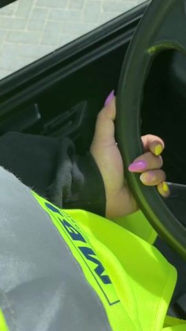 A first-person perspective video shows someone operating a Linde forklift truck on what appears to be an industrial site. The driver, wearing a yellow safety vest, has distinctive purple and yellow nails. The video shows the truck's dashboard with various controls, a small display screen, and a steering wheel bearing the Linde logo. The driver is steering the vehicle on a paved area alongside a chain-link fence. A reflection in the rearview mirror shows another forklift in the background.