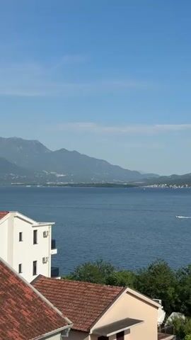 The video shows a daytime view of a body of water, likely a bay or a wide river, from an elevated perspective. In the foreground are rooftops of buildings with red clay tiles, interspersed with trees. The body of water fills most of the frame, with land visible in the distance. Mountains form a backdrop to the landscape on the far side of the water. The sky is blue with some wispy white clouds. Two boats are seen speeding across the water, creating white wakes behind them.  The video appears to be stable and the lighting is bright, giving a clear view of the scene.