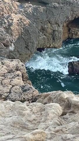 The video shows a choppy ocean with white waves crashing against rocky formations. The water is a deep blue-green color. The rocks are rough and varied in texture, with some appearing porous and others smoother. Sunlight glints off the water, creating highlights and shadows on the rocks. The scene appears to be a natural coastal environment, likely a cove or inlet. There are no people or man-made objects visible in the frame. The camera angle is fixed, looking down at the water and rocks. The video is brief, showing only a few seconds of the wave action.