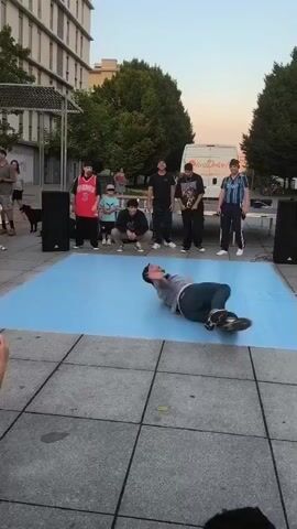 The video shows a breakdancer performing on a blue mat in an outdoor public space. They are wearing a grey t-shirt, blue jeans, and white sneakers. The dancer is in the middle of a complex move, with their body contorted and legs in the air, supported by one hand on the mat. Several spectators are gathered around the mat, some seated on the ground and others standing. Behind the dancers, there are buildings, trees, and a white van. The sun appears to be setting, casting a warm light on the scene.