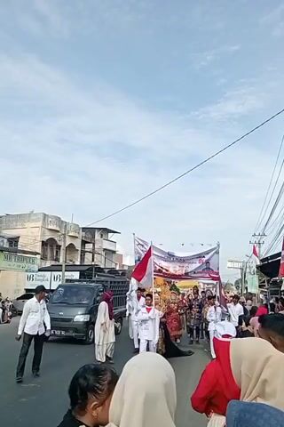 A parade is taking place on a street lined with buildings. A black pickup truck is parked on the left side of the street. A group of people are walking in a procession, some carrying flags and a banner. One person in the center of the procession is wearing traditional clothing and a headdress. Onlookers are standing on the side of the street watching the parade. The sky is cloudy.