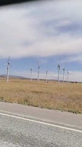 The video shows a landscape with several wind turbines visible in the distance. The turbines are white and have blades that appear to be slowly rotating. They are situated in a grassy, somewhat dry field. In the foreground, a grey road surface is visible, suggesting the shot was taken from a moving vehicle. The sky is blue with scattered white clouds. The overall impression is a rural or semi-rural setting with renewable energy infrastructure.