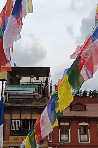 The video shows a string of colorful prayer flags fluttering in the wind against a cloudy sky. The flags are strung across a street, passing in front of traditional buildings. One building is painted red and has several windows with white curtains. Another building to the left appears to have a rooftop seating area. The prayer flags are made of various colors, including blue, white, red, green, and yellow, and appear to have printed text on them. The overall impression is one of a vibrant, possibly cultural or spiritual, setting.
