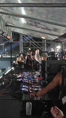 The video shows a DJ performing at a large outdoor music festival at night. The DJ is standing behind a mixing console, gesturing with his hands, with a small group of people looking on from behind. The video then quickly pans to the crowd, showing a large mass of people with their hands raised, illuminated by stage lights. The crowd is dense, with people holding phones and recording the performance. The environment is an open outdoor space, surrounded by buildings and trees. A radio tower is visible on the horizon.