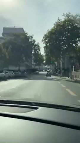 The video shows a car driving down a city street. The street is lined with buildings and trees. There are parked cars on both sides of the street. Pedestrians are walking on the sidewalks. A church can be seen on the right side of the street. The sky is bright, and the sun is shining.