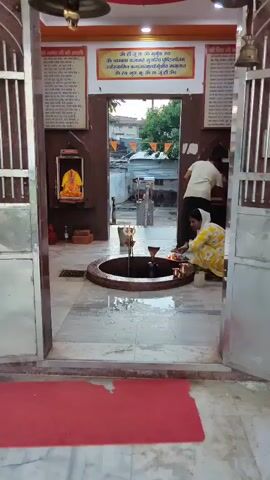 This video shows a religious ceremony taking place inside a temple. The focus is on a large, dark, round structure in the center of the frame, which appears to be a Shiva Lingam, a sacred idol in Hinduism. A man in a light-colored shirt and dark pants is pouring water from a bucket onto the Shiva Lingam using a ladle. Next to him, a woman wearing a yellow and white patterned outfit and a white head covering is performing a ritual, possibly offering prayers or placing items. She is holding a small plate with offerings. Both individuals are barefoot, indicating respect within the sacred space. The floor is wet and appears to be made of marble. In the background, through an open doorway, glimpses of the outside world can be seen, including some buildings and flags. The temple interior is decorated with religious imagery and signage, suggesting a devotional atmosphere.