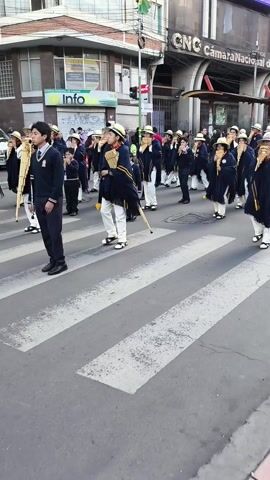 A group of people wearing matching outfits and playing panpipes are walking in a parade across a crosswalk on a city street. The people are wearing white pants, sandals, navy blue ponchos with yellow stripes, and light colored hats. The group is primarily in the crosswalk, but some people are still on the curb behind them. On the right side of the street is a building with the words "Cámara Nacional de Comercio" on it. On the left is a building with a sign that says "Info Agencia Municipal de Turismo". The parade appears to be taking place during the daytime. There are other people standing and watching the parade along the sides of the street.