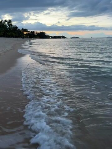 This media file captures a serene beach scene at Baan Mae Nam, Thailand, during dusk. The primary subjects are the gentle waves lapping against a sandy shore and the vast expanse of the ocean. In the distance, a line of palm trees and some structures suggest a coastal town or resort area. A couple of indistinct figures can be seen walking along the beach in the mid-ground, adding a human element to the tranquil landscape.

The time of day is clearly evening, with the sky displaying a dramatic interplay of dark, moody clouds tinged with the warm hues of sunset – oranges and yellows peeking through. This creates a soft, diffused light over the scene. The weather appears calm, with the sea surface rippling gently rather than showing any signs of rough conditions.

The location cues, like the tropical vegetation and the calm, shallow water, are consistent with a Southeast Asian beach setting, further supported by the provided location context of Baan Mae Nam, Thailand. The overall mood evoked is one of peace, tranquility, and the quiet beauty of nature at the end of the day. There is no visible text in the media file.