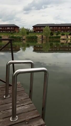 The scene is a tranquil, overcast day at a lakeside resort. The camera, positioned on a wooden dock, focuses on the stainless steel ladder leading into the calm, reflective water. In the background, two multi-story, dark-wood buildings are visible, their reflections mirrored in the lake's surface.  A line of green shrubbery and small trees separates the buildings from the water's edge. The sky is filled with grey clouds. The calm water and mirrored reflections create a sense of peace and serenity. A child's voice can be heard saying, "Let's go after the roof."