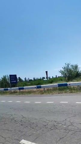 The video captures a moving shot from a vehicle, showing a roadside scene on a clear, sunny day. In the foreground, a paved road with white dashed lines is visible. A white and blue metal guardrail runs parallel to the road, slightly above the road level. Beyond the guardrail, a line of tall, slender trees, possibly poplars, extends into the distance. At approximately 0:03, a blue square road sign with a white arrow indicating a turn is visible among the trees. The sky is bright blue and cloudless. The camera maintains a consistent forward movement throughout the clip.