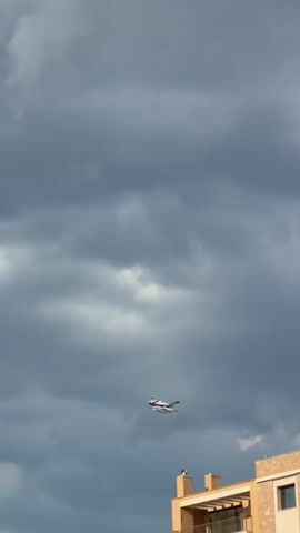 A small airplane flies across a cloudy sky above a row of buildings. The plane is white with a red stripe and is moving from left to right. The buildings are a mix of colors, including beige, orange, and brown, and have balconies and windows. A streetlamp is visible on the right side of the frame. The sky is filled with dark, dramatic clouds, with a small patch of blue sky visible in the upper right corner. The overall impression is one of an approaching storm.