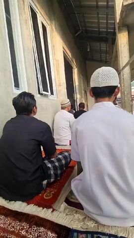 Here is a concise description of the video:

The video shows a group of men sitting on prayer rugs, presumably engaged in a religious activity, such as prayer or a sermon. They are positioned outdoors, along the side of a building with a window, under a partially covered area. The men are wearing traditional Islamic clothing, including white caps, and are facing in the same direction. The environment is a simple, possibly residential or community area.