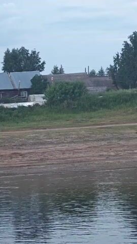 This video shows a view of a riverside village in Vorogovo, Russia. The scene is dominated by a body of water in the foreground, with a dirt bank leading up to a grassy slope where several wooden houses are situated. The houses appear to be older, some with weathered wood exteriors and simple, pitched roofs. Trees are scattered throughout the landscape, some of which are quite large and leafy, suggesting a rural or semi-rural setting. The sky is overcast with some light clouds, indicating a possibly mild or cool day. The time of day appears to be either early morning or late afternoon due to the diffused light. There is no visible text in the video. The overall mood is tranquil and somewhat rustic, capturing a glimpse of everyday life in a Russian village. The activity taking place is not explicitly shown, but the scene suggests a static view of the village from across the water, possibly from a boat or the opposite bank.