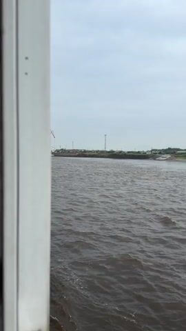 The video shows a river scene from the perspective of a moving boat, looking towards the far bank under an overcast sky during the daytime. The camera pans from left to right, revealing various elements along the riverbank.

On the far bank, industrial infrastructure is visible. Initially, a large red and white industrial crane stands prominent, with what appears to be a vessel docked nearby a concrete structure. As the camera pans, a tall metal tower comes into view further along the bank. The bank itself features extensive concrete retaining walls and large piles of rocks or gravel, suggesting a port or industrial loading area. Green foliage and trees are interspersed along the bank. The river water in the foreground is dark and choppy, indicating movement of the boat or significant currents.

The scene is consistent with a port or industrial area along a river, such as Nizhneleninskoe, Russia. No visible text is present in the video.