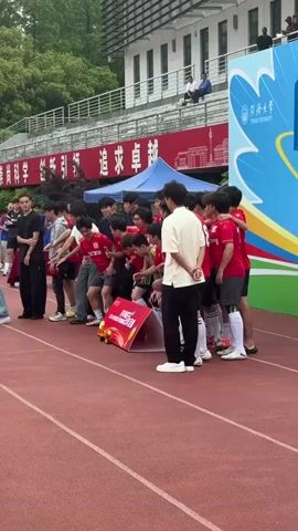 A group of approximately 15 young men on a track embrace and extend their arms out, cheering. They are clustered around a low, red and gold sign on the ground that appears to indicate a win. Some are wearing red soccer uniforms, and others casual clothing. In front of the group are two men in casual clothing, but more formal than the young men. Behind the group is a large blue, white, and yellow graphic with red Chinese script in the background. The stadium is visible with people sitting on the upper level. There are other individuals also present on the track.