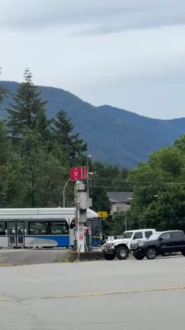A bus is stopped at a gas station. A white Jeep is parked to the right of the bus. A dark SUV is parked behind the Jeep. A tall, slender sign with red and white lettering is to the right of the bus. Trees and mountains are in the background.