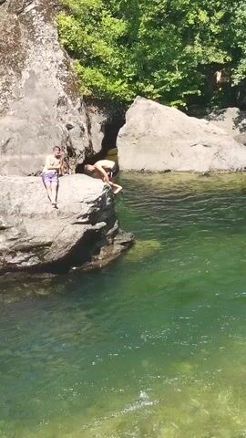 Two young men are on a large rock formation near a body of water. The man on the left is standing on the edge of the rock, facing the water. The man on the right is bent over, appearing to be preparing to jump. The water is clear and green. Trees and other rock formations are visible in the background. The sky is blue.