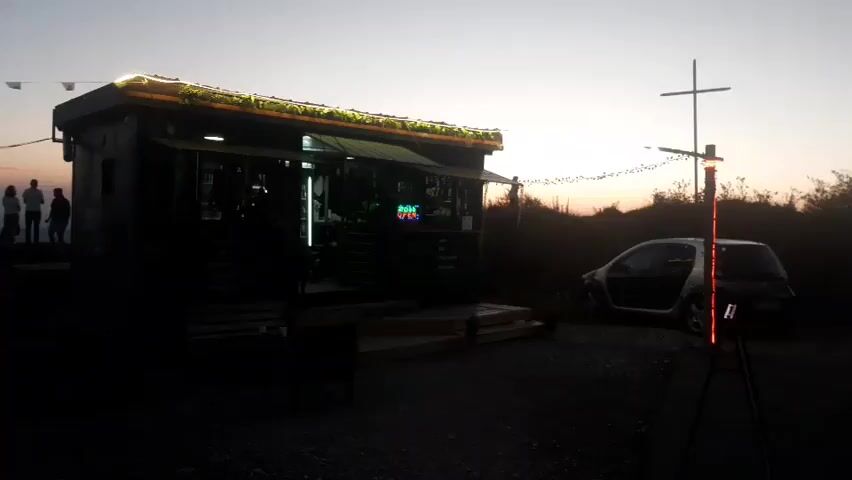 The video begins with a wide shot of a small, rustic stand or hut situated outdoors as dusk settles. The structure has an awning with decorative lights strung along its edge. A small smart car with its door open is parked to the right of the stand. In the foreground, a tripod with a vertically oriented light fixture that has red and orange lights is visible. To the left of the stand, a group of silhouetted figures stand on a slight incline, facing away from the camera. The background consists of a darkening sky and silhouetted foliage. The overall impression is of a remote or secluded location, possibly a roadside stop or a small business operating in an outdoor setting at twilight. The scene is lit by the lights on the stand and the light fixture on the tripod, with ambient twilight providing the backdrop.