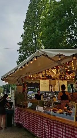 This media file depicts an outdoor food stall, likely at a fair or market, set against a backdrop of lush green trees and a bright, possibly overcast, sky. The stall is adorned with numerous string lights, suggesting it might be late afternoon or early evening. The main subject is the food stall itself, a covered structure with a checkered red and white tablecloth draped over its front. Display cases in front of the stall showcase an array of food items, including what appears to be pastries and prepared dishes.

Several people are visible interacting with the stall. Two individuals, a man and a woman, are working behind the counter, presumably serving customers. A group of customers, including a couple in the foreground, are looking at the offerings and seemingly deciding what to order. One customer, a woman with dark hair, is leaning her head on the shoulder of the man in front of her, suggesting a relaxed and perhaps intimate moment. Another customer, a man with gray hair and a pink shirt, is looking upwards, possibly at the menu or the lights. Further back, other patrons can be seen, adding to the bustling atmosphere of the scene.

A digital menu board is prominently displayed on a stand in front of the stall. While the text on the menu board is not clearly legible, it indicates that information about the food items is available. The overall scene conveys a sense of enjoyment and casual dining, typical of a public gathering or event. The presence of large trees suggests an outdoor park or recreational area. The weather appears to be fair, with a soft, diffused light illuminating the scene.