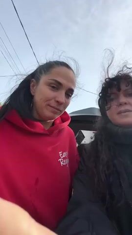 Two women are filmed outdoors in what appears to be a rural setting. The woman on the left, wearing a red hooded sweatshirt with "East Region" written on it, has her hair pulled back in a ponytail. The woman on the right, wearing a black jacket and a dark turtleneck, has long, dark curly hair. The woman on the right is speaking and gesturing, while the woman on the left looks at her and then smiles and waves at the camera. In the background, there are houses, a blue fence, a car, and some electrical wires. A glass of red liquid is visible, held by the woman on the left at one point. The sky is visible and appears to be partly cloudy.