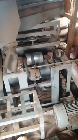The video shows a close-up view of an industrial machine, likely a separator or sorter. The machine appears to be made of metal and has several components, including a hopper at the top, a series of sieves or screens, and a rotating drum or cylinder. There is also an electric motor attached to the machine. The environment appears to be a workshop or factory setting, with sacks of material visible in the background. The machine is either in operation or has recently been used, as indicated by dust and debris on its surfaces. The video is shaky and the lighting is dim, making it difficult to discern fine details.