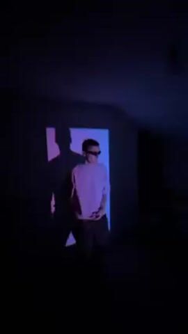 The video depicts a person wearing sunglasses and a light-colored t-shirt in a dark room. The person is standing in front of a rectangular red light that is also casting a shadow. The person poses, puts their arms on their chest, and dances with casual hand movements in front of the red light.