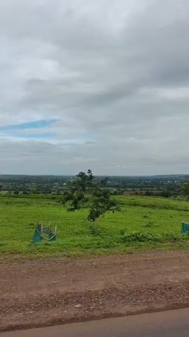 The video is a panoramic shot of a green landscape under a cloudy sky. The camera pans from left to right, revealing rolling hills covered in lush green grass and scattered trees. In the foreground, there is a dirt path or road. In the distance, a small town or village can be seen nestled among the hills. The overall impression is one of a serene and natural environment.
