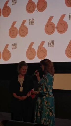 Two women stand on a stage in front of a screen. The screen displays a repeating logo of a number 66 incorporating different images. The logo reads "Zlín Film Festival". The woman on the left has short light brown hair, is wearing a black dress or jacket, a white blouse, and a name tag. The woman on the right has shoulder-length brown hair and is wearing a blue dress with a botanical print. She holds a red program and a microphone, and speaks into the microphone while gesturing with her hands. She occasionally glances at the woman next to her. The environment appears to be a movie theater, with rows of seats visible in the foreground.