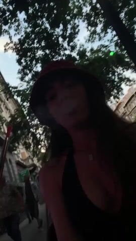A woman wearing a red hat and dark clothing is in the foreground, exhaling a large cloud of smoke or vapor. She is looking to her left, and her face is obscured by the smoke. In the background, there are people walking on a sidewalk, trees, and buildings. The lighting suggests it is daytime, possibly late afternoon or early evening.