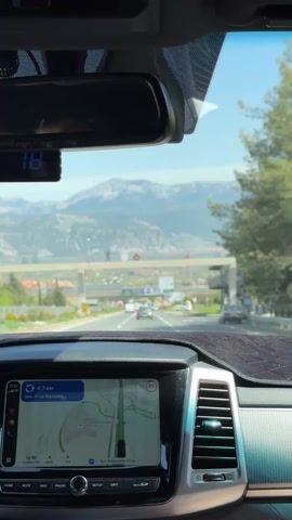 The video shows a car driving on a highway toward mountains. The dashboard indicates a navigation system is running, showing a route 4.6 km ahead. The outside temperature display reads 16 degrees Celsius. Other cars are visible on the highway. The scene is shot from inside the car, looking out through the windshield.