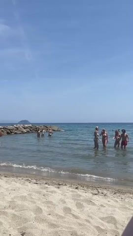 The video shows a beach scene on a sunny day with a blue sky. Several people are standing in the water, some close to the shore and others further out, near a rock barrier. The people are dressed in swimwear. The water is calm, with small waves lapping on the sandy beach. In the foreground, the sand is a light tan color. The overall scene suggests a warm, leisurely atmosphere.