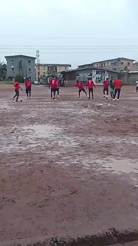 A group of young men in red shirts and black shorts are practicing soccer on a muddy field. The field is covered in puddles and dirt. In the background, there are several buildings, including a tall apartment building and a smaller, more run-down structure. The sky is overcast. The players are spread out across the field, some with soccer balls at their feet. One player in a green shirt and black shorts appears to be practicing separately from the main group. The overall scene suggests an informal soccer practice session taking place in less than ideal weather conditions.