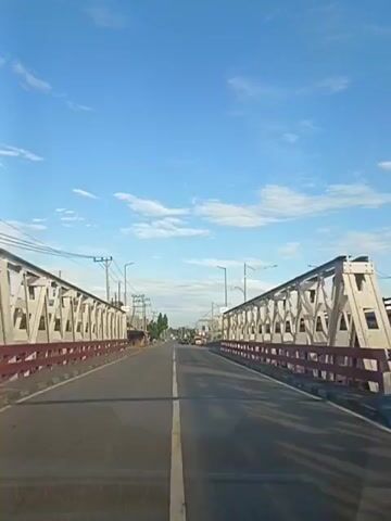 The video shows a forward-facing view from a vehicle traveling on a paved road. The road is flanked on both sides by metal bridges with white and red railings. The sky is blue with scattered white clouds. Along the sides of the road, there are buildings, power lines, and streetlights. Some vegetation is also visible. The video appears to be captured during the daytime, possibly in the morning or late afternoon, given the lighting conditions. There are no people or other vehicles clearly visible in the immediate vicinity, but there are suggestions of distant traffic.