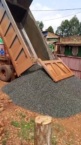 A brown dump truck is raising its cargo bay to dump a load of small gravel onto a patch of reddish-brown dirt. The truck bed is dirty and appears to have a wooden panel across its bottom. The gravel forms a large, conical pile. A man in a dark jacket, jeans, and a face mask stands to the side, watching the gravel being dumped. In the background, there are low buildings with corrugated metal roofs and fences. There are also utility poles and trees visible in the distance. The ground is uneven, with scattered rocks and patches of grass. The sky is overcast.