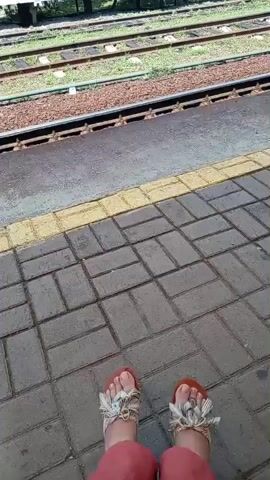 The video shows a pair of feet wearing sandals, standing on a brick-paved platform next to train tracks. The feet are positioned in the center of the frame, with the toes pointing slightly inward. The sandals are open-toed with decorative embellishments. The platform is wet, suggesting recent rain. The train tracks are visible in the background, with ballast and grass. The overall scene appears to be at a train station.