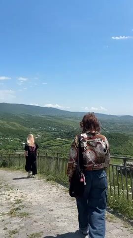 The video shows a panoramic view of a lush green valley with scattered houses and rolling hills under a clear blue sky with some white clouds. Two women are walking away from the camera on a paved path with a metal railing to their right. The woman in the foreground is wearing a patterned shirt and blue jeans, and has a black bag slung over her shoulder. The woman in the background has blonde hair, is wearing a black t-shirt and black pants, and has a black bag. The path appears to be sloped, leading downwards into the valley.