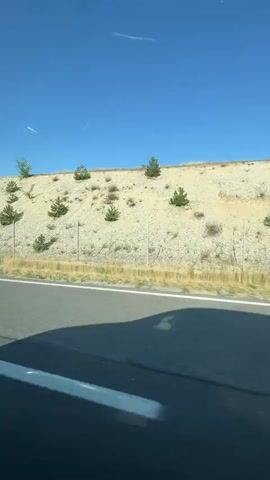 The video shows a view from inside a car traveling on a highway. The car is moving forward, and the landscape outside consists of a sandy, hilly area with sparse vegetation and some trees. The sky is clear and blue. The interior of the car is visible, including the dashboard, a side mirror, and the car's interior trim. The road ahead is visible, with a white line marking the lane. The video appears to be a first-person perspective of a passenger or driver.