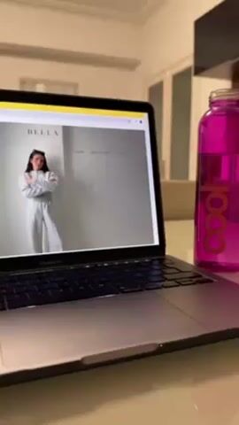 A close-up video shows a laptop on a table with a pink water bottle beside it. The laptop screen displays a website for a clothing brand called BELLA. A model is shown wearing a light gray tracksuit. The camera zooms in on the model's face. The laptop is a Macbook Pro. The background is a room interior with a white door and a neutral-colored wall.