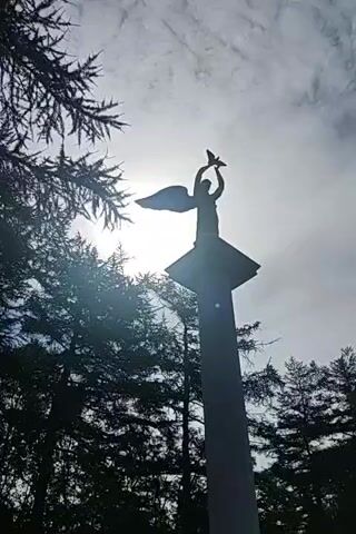 A low-angle video shows a white monument topped with a silhouetted angel holding a bird. The monument is surrounded by trees, and the sun shines brightly behind the angel, creating a silhouette effect. The sky is partly cloudy. The video pans slightly upwards as it progresses.