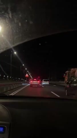 The video shows a view from inside a car driving on a highway at night. The road is lit by streetlights and the headlights and taillights of other vehicles. Traffic appears moderate, with several cars and a large truck visible ahead. A triangular road sign with an image of a person falling is visible on the left side of the road. The dashboard of the car shows a digital display, but the numbers are partially obscured. The scene suggests a typical highway commute or travel at night in a developed area. There is no explicit indication of weather conditions, but the darkness implies it is not daytime. The location is identified as Azor, Israel. No specific emotions are conveyed through the visual content, which remains objective and descriptive of the scene.