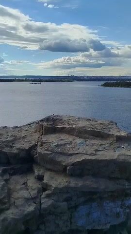 A drone shot looks down from a rocky cliff edge over a wide, calm blue river. The river has a slight ripple. In the distance, the opposite bank of the river is visible, with a thin line of green trees and a distant city skyline under a bright blue sky with scattered white clouds. A small motorboat is moving across the water, leaving a white wake.