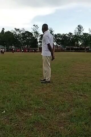 The video shows a man standing on a grassy field, with his hands clasped behind his back. He is wearing a white short-sleeved shirt and light-colored pants. In the background, there is a volleyball net and people appearing to play volleyball. Trees border the field in the distance. The sky is overcast.