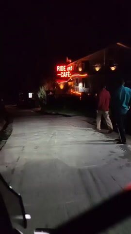 The video shows a nighttime scene from the perspective of someone on a vehicle, likely a motorcycle given the handlebars visible in the foreground. The camera is pointed down a paved road that is illuminated by headlights from the vehicle. To the right of the road, there is a building with a neon sign that reads "RIDE INN CAFE." The building is lit up with warm-toned lights, and there appears to be some outdoor seating. A person in a maroon top and light-colored pants is walking away from the camera on the sidewalk in front of the cafe. There are trees and foliage on either side of the road and around the cafe. The overall impression is a dimly lit, somewhat rural or small-town setting at night.