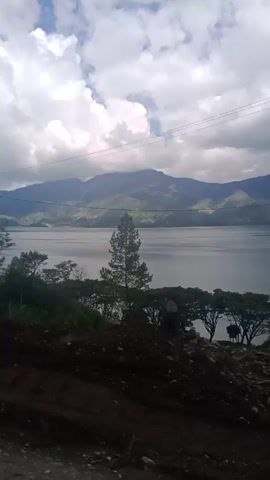 The video shows a landscape view, likely from a moving vehicle, featuring a body of water bordered by mountains and trees. The sky is partly cloudy. The foreground consists of rough terrain with dirt and rocks. In the middle ground, there are trees, including pine trees, and vegetation lining the shore of the body of water. The background displays rolling hills and mountains, with some areas shrouded in mist or clouds. Power lines are visible in the upper portion of the frame. The camera angle is slightly tilted, giving a sense of looking down and across the scene. The lighting suggests it is daytime, possibly overcast.