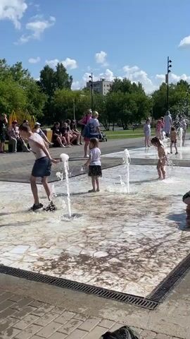 A group of children and adults are gathered in an outdoor splash pad area on a sunny day. Water jets spray upwards from the tiled ground, creating a fun and refreshing environment for the children who are actively playing and running through the water. Some children are wearing swimwear, while others are in regular clothing. Adults are observing from the sidelines, some seated on benches in the background. The area is surrounded by trees and grass, indicating a park or public recreational space. The overall atmosphere appears joyful and active, with the focus on the children's enjoyment of the water features.
